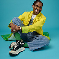 WBM is smiling and sitting on the floor while wearing a bright yellow and green jacket, overalls and black sneakers in front of a blue ba...